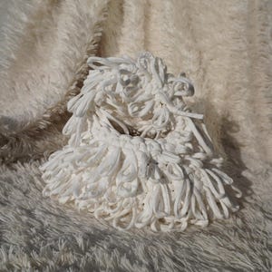 May include: A white, handmade, circular basket made of looped fabric. The basket has a textured, fluffy appearance and sits on a soft, white, faux fur surface. The basket's design features a woven base with looped fabric extending outwards.