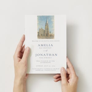 Könnte beinhalten: Eine Hochzeitskarte, die von Händen gehalten wird und eine Aquarellillustration einer Kirche zeigt. Die Einladung enthält die Namen Amelia und Jonathan sowie Datum und Uhrzeit der Hochzeit. Der Text ist dunkelblau.