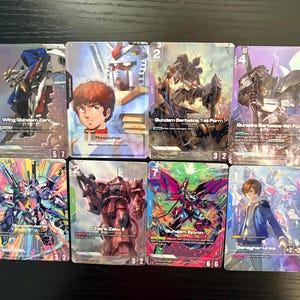 May include: Eight trading cards featuring Gundam characters and mechs. The cards display colorful illustrations of robots and anime characters. The cards have various numbers and text, including "Wing Gundam Zero", "Gundam Barbatos 1st Form", and "Char's Zaku II".