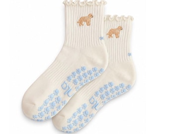 May include: A pair of cream-colored ribbed socks with ruffled cuffs. Each sock features a small embroidered golden-brown dog and light blue silicone paw print grips on the sole. The text "pu" is visible in light blue on the side of each sock.