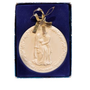 May include: Cream-colored ceramic Christmas ornament featuring a relief of a bride and groom embracing. The ornament is round, with floral detailing and the inscription "Our First Christmas Together." It is presented in a blue box with a gold ribbon.