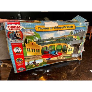 May include: A Thomas & Friends TrackMaster Thomas at Tidmouth Sheds railway system toy set in a rectangular cardboard box. The box features blue and yellow graphics, a depiction of the train shed, a blue locomotive, and grey duct tape.