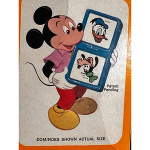 May include: Vintage Mickey Mouse dominoes set featuring Mickey Mouse holding a blue dominoes game with Donald Duck and Goofy. The dominoes are shown actual size. The text "Patent Pending" is visible. The packaging is orange and white.