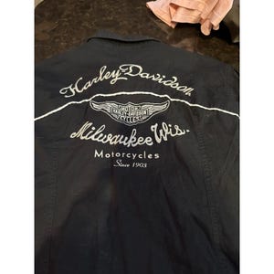 Może przedstawiać: Czarna koszula zapinana na guziki z białym haftowanym logo i tekstem Harley-Davidson. Logo zawiera skrzydlaty emblemat i słowa "Motor Harley-Davidson Cycles". Tekst brzmi "Milwaukee Wis. Motorcycles Since 1903."