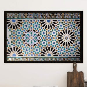 May include: A framed mosaic with a black border, featuring a repeating pattern of blue, black, and white floral designs. The artwork hangs above a wooden kitchen counter with utensils.