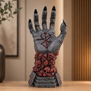 Peut inclure: Une sculpture grise et rouge représentant une main avec des ongles noirs, un symbole rouge sur la paume et une base de visages et de crânes rouges. La sculpture est sur une surface en bois.
