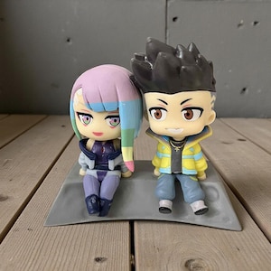 May include: Two small figurines of anime-style characters. One has purple and blue hair, the other has black spiky hair and a yellow jacket. They are sitting on a gray platform, with a gray wall in the background.