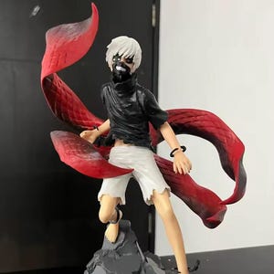 May include: A detailed figurine of a character with white hair, a black mask, and a black outfit, paired with white shorts. The figure has large, flowing red appendages and is posed on a gray, rocky base.