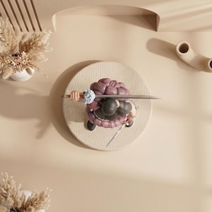May include: A figurine of a character with a purple brain-shaped head, black clothing, and a sword, positioned on a circular, textured beige surface. The figure is holding a sword and has a chain around its waist. The background is a light beige color.