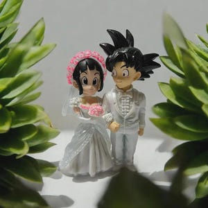 以下が含まれることがあります： アニメスタイルの2体のフィギュアが特徴のウェディングケーキトッパー。花嫁は白いガウン、ベール、ピンクの花のヘッドピースを着用し、ブーケを持っています。新郎は白いスーツを着ています。フィギュアは白い背景に配置され、緑の葉で縁取られています。