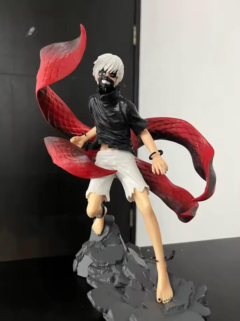 May include: A detailed figurine of a character with white hair, a black mask, and a black outfit, paired with white shorts. The figure has large, flowing red appendages and is posed on a gray, rocky base.