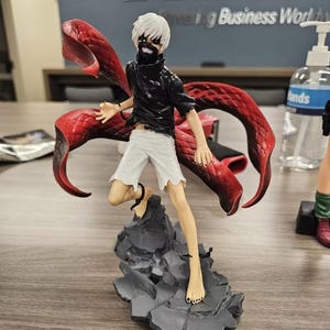 May include: A detailed figurine of a character with white hair, a black top, and white shorts, standing on a gray rocky base. The figure has a red, wing-like structure extending from its back. The figure is barefoot and has a masked face.