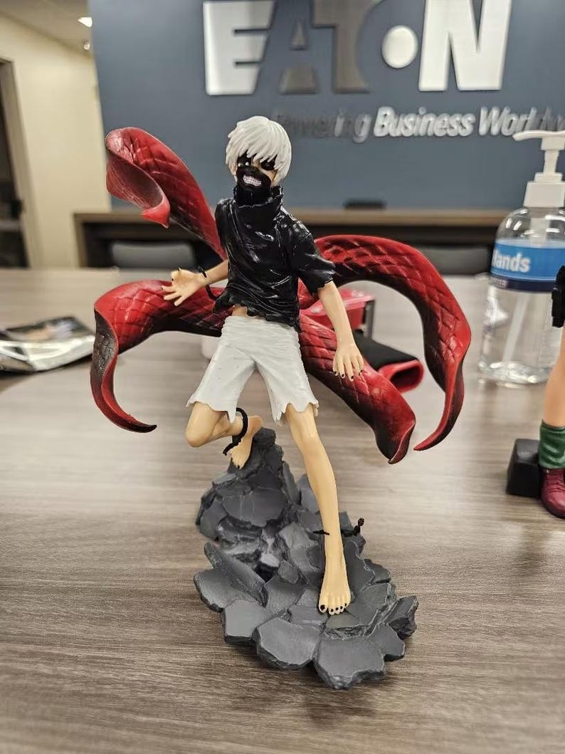 May include: A detailed figurine of a character with white hair, a black top, and white shorts, standing on a gray rocky base. The figure has a red, wing-like structure extending from its back. The figure is barefoot and has a masked face.