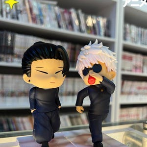 Puede incluir: Dos figuras de estilo anime. Una tiene cabello negro y viste un atuendo negro. La otra tiene cabello blanco, gafas de sol y un atuendo negro. Las figuras est&aacute;n frente a una estanter&iacute;a llena de libros.