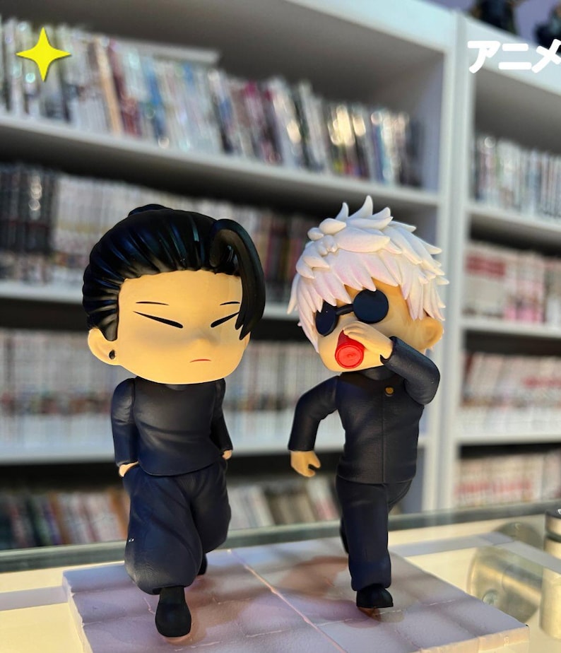 Puede incluir: Dos figuras de estilo anime. Una tiene cabello negro y viste un atuendo negro. La otra tiene cabello blanco, gafas de sol y un atuendo negro. Las figuras est&aacute;n frente a una estanter&iacute;a llena de libros.