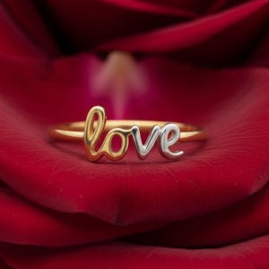 May include: A gold and silver ring with the word "love" in a cursive font. The ring is set against a red rose, creating a romantic and elegant aesthetic. The ring's design features a gold band and silver lettering.