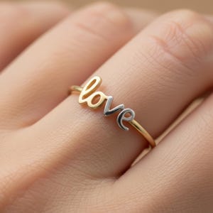 May include: A gold and silver ring spelling out the word "love" in a cursive font. The ring is a delicate piece of jewelry, with the word "love" in gold and silver tones. The ring is on a finger.