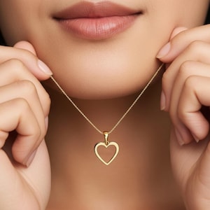 May include: A gold-colored heart pendant necklace. The heart-shaped pendant is hollow and hangs from a delicate chain. The necklace is being held up by a person's hands.