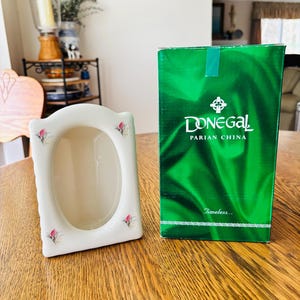 May include: A white ceramic picture frame with a floral design and a green box with the text "Donegal Parian China". The frame has an oval opening and is decorated with pink flowers. The box has the words "Timeless..." printed on it.