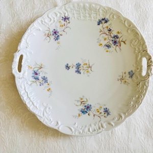 May include: A round white porcelain serving platter with two integrated handles. The rim features an embossed scrollwork pattern. The center and edges are decorated with small, delicate floral sprays in shades of blue and yellow.