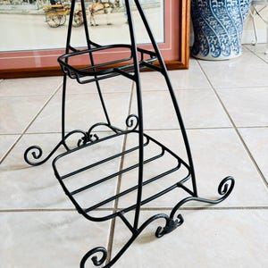 May include: A black metal two-tier display stand featuring a triangular frame with a top shelf and a larger bottom shelf. The structure is supported by four curved, scroll-style feet at the base, providing a stable foundation for the tiered shelving.