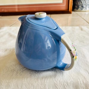 May include: A light blue ceramic teapot with a matching lid and a white handle adorned with floral patterns. The teapot has a rounded body and a spout for pouring. The lid features a small, round knob with a floral design.