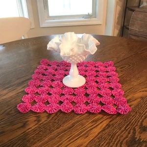May include: A white milk glass candy dish with a ruffled edge sits on a pink crocheted doily on a wooden table.