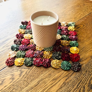 May include: A cylindrical candle in a light pink ceramic vessel with a textured diagonal line pattern, resting on a square mat constructed from numerous small, gathered fabric circles in shades of red, green, yellow, and dark brown.