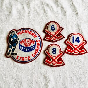 May include: A collection of four embroidered patches including one large circular patch featuring a hockey player and the text Michigan Pee Wee AA State Champs 1978-79, alongside three smaller shield-shaped patches with numbers 6, 8, and 14.