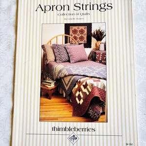 May include: A book cover titled "Apron Strings Collection of Quilts" by Lynette Jensen. The cover features a photograph of a bed with a quilt, pillows, and a small wooden table. The quilt is a mix of burgundy, brown, and cream colors.