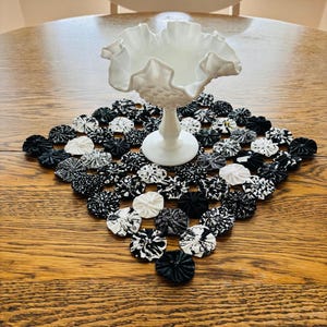 May include: A white milk glass pedestal bowl with a ruffled rim sits atop a square fabric mat. The mat is constructed from numerous small, circular fabric yo-yos in various black and white patterns, including solids, stripes, and floral prints.