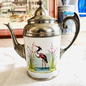 May include: A vintage white porcelain teapot with a silver-toned lid, handle, and spout. The teapot features a hand-painted scene of a bird standing in water with reeds. The teapot is a decorative item.