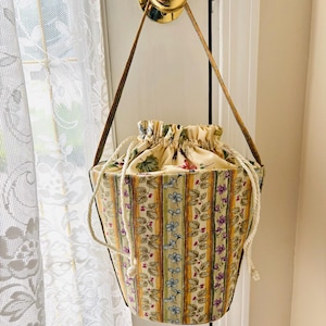 May include: A fabric bucket bag with a drawstring closure, featuring a vertical striped pattern in yellow, green, and cream, decorated with small floral and berry motifs. The bag includes a long, thin brown strap for hanging or carrying.