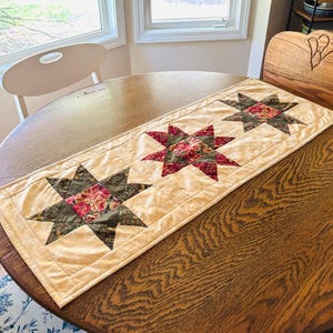May include: A quilted table runner with three large star patterns. The stars feature a floral print in red and pink, set against a green and tan background. The runner has a cream-colored border and is placed on a wooden table.