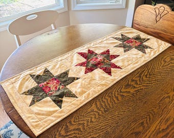 May include: A quilted table runner with three large star patterns. The stars feature a floral print in red and pink, set against a green and tan background. The runner has a cream-colored border and is placed on a wooden table.