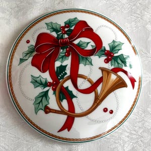 May include: A white ceramic plate with a Christmas design. The plate features a red bow with green holly leaves and red berries, and a gold-colored horn. The plate is surrounded by a brown rope border.