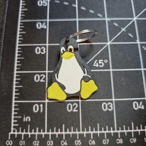 May include: A black, white, and yellow penguin keychain featuring a stylized character design. The keychain is positioned on a black cutting mat with white grid lines and inch markings, showing the item is approximately 1.5 inches in height.
