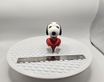 May include: A Snoopy figurine with a red heart-shaped body and white feet. The black and white figure is sitting on a white surface. A metal ruler is placed in front of the figurine.
