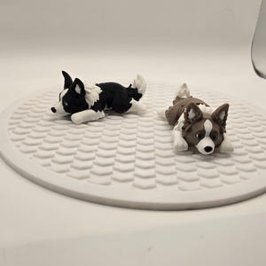 May include: Two miniature dog figurines, one black and white, the other brown and white, are lying down on a white, textured, circular surface. The dogs are detailed with realistic features, creating a charming display.