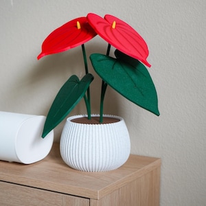 May include: A decorative artificial anthurium plant in a white, ribbed ceramic pot. The plant features vibrant red flowers with yellow centers and dark green leaves. The pot sits on a light wood surface.
