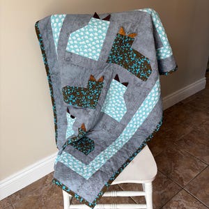 May include: A handmade quilt draped over a white chair. The quilt features a grey background with large, stylised horse head shapes in teal and brown. The quilt's border is brown with a teal and brown pattern.