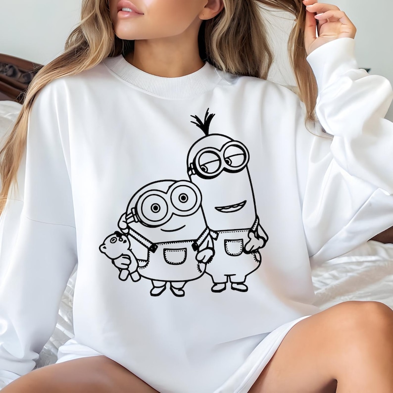 May include: White sweatshirt with a black graphic of two Minions. One Minion holds a teddy bear. The Minions are wearing dungarees and goggles. The sweatshirt has a crew neck.