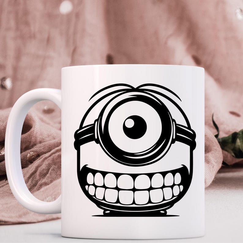 May include: White ceramic mug with a black and white cartoon Minion design. The illustration shows a single eye, a wide grin, and goggles. The mug has a curved handle. The design is a popular character from a children's film.