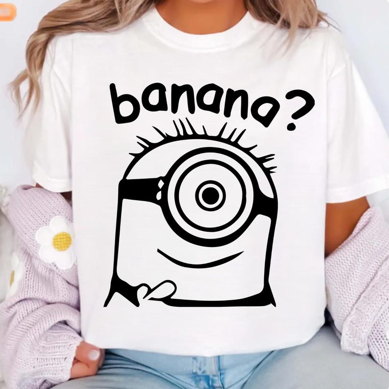 May include: White t-shirt with a black cartoon Minion graphic, including one eye, goggles, and the question "banana?" above. The design is a fun, cartoon style.