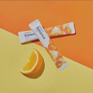May include: Two white rectangular Unicity Balance Orange supplement stick packs featuring a geometric orange circle pattern. Each pack displays the text Unicity Balance Orange and a net weight of 7.25 g. A fresh orange wedge is positioned nearby.