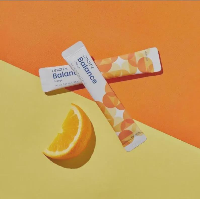 May include: Two white rectangular Unicity Balance Orange supplement stick packs featuring a geometric orange circle pattern. Each pack displays the text Unicity Balance Orange and a net weight of 7.25 g. A fresh orange wedge is positioned nearby.