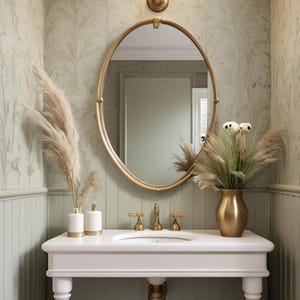 May include: A bathroom vanity with a white countertop and a gold-framed oval mirror. The sink has gold fixtures. Decorative vases with dried plants and a wall-mounted light fixture complete the elegant design. The walls are painted in a light green color.