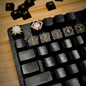 May include: Black mechanical keyboard with custom keycaps. The keycaps have gold accents and symbols, including numbers and stylized characters. Some keycaps feature unique designs, such as a sun and a face. The keyboard is on a wooden surface.