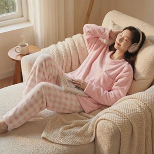 May include: A person wearing pink pajamas and headphones relaxes on a cream-colored chaise lounge. The pajamas include a long-sleeved top and checkered pants. A book rests on the person's lap, and a cup of coffee sits on a nearby table.