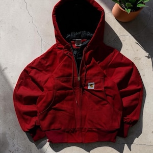 May include: A red zip-up hooded jacket with a black interior lining and a drawstring hood. The jacket features a small rectangular Carhartt brand patch on the front left chest pocket area and ribbed cuffs and hem for a secure fit.